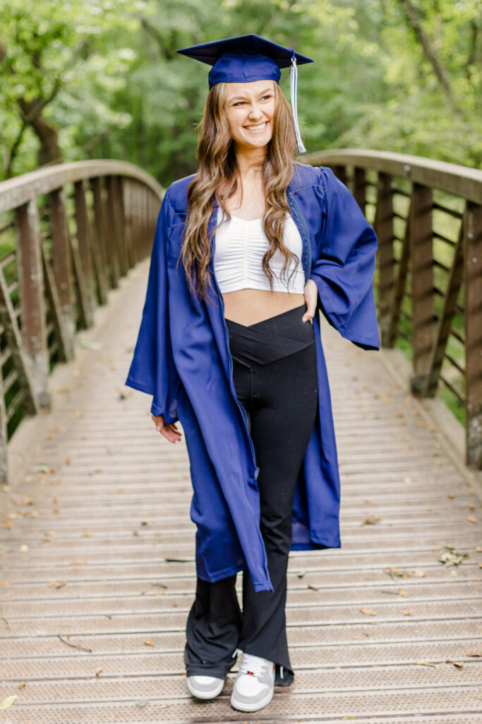 Senior girl wearing a cap and gown walks across the old bridge at Williams Mill Greenspace in Flowery Branch, Georgia