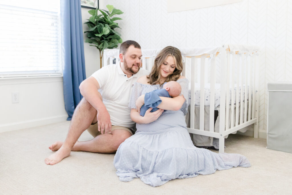 A family sits in their nursery holding their newborn baby boy and smile down at him at a lifestyle newborn session.