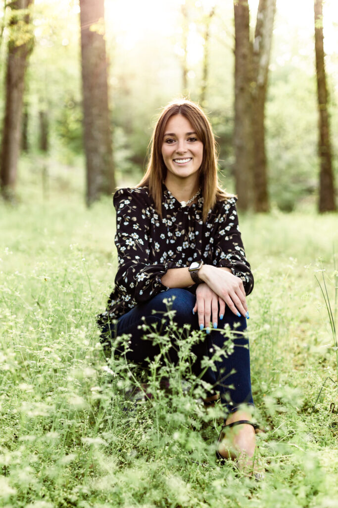 Senior girl sits in a meadow in front of trees in Flowery Branch, Georgia
