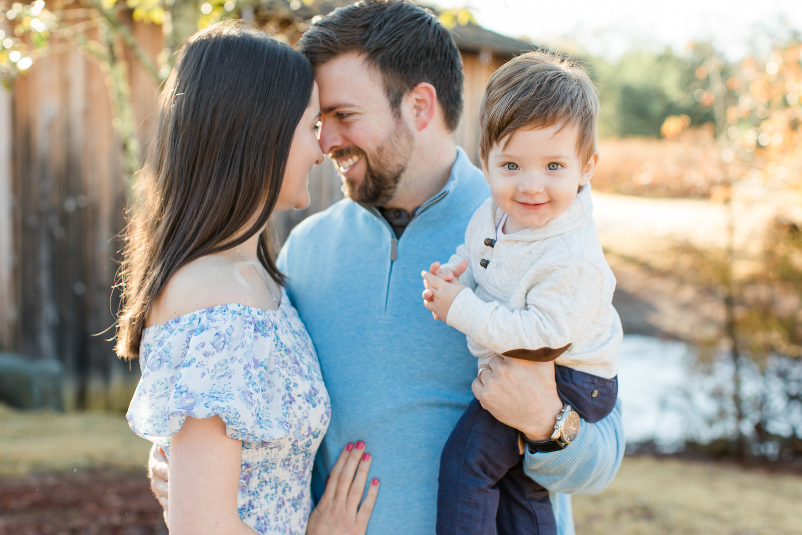 Parents hold toddler son in arms and look at each other. Boy looks at camera and smiles. They wear light coordinating colors for their photo session in Roswell, Georgia.
