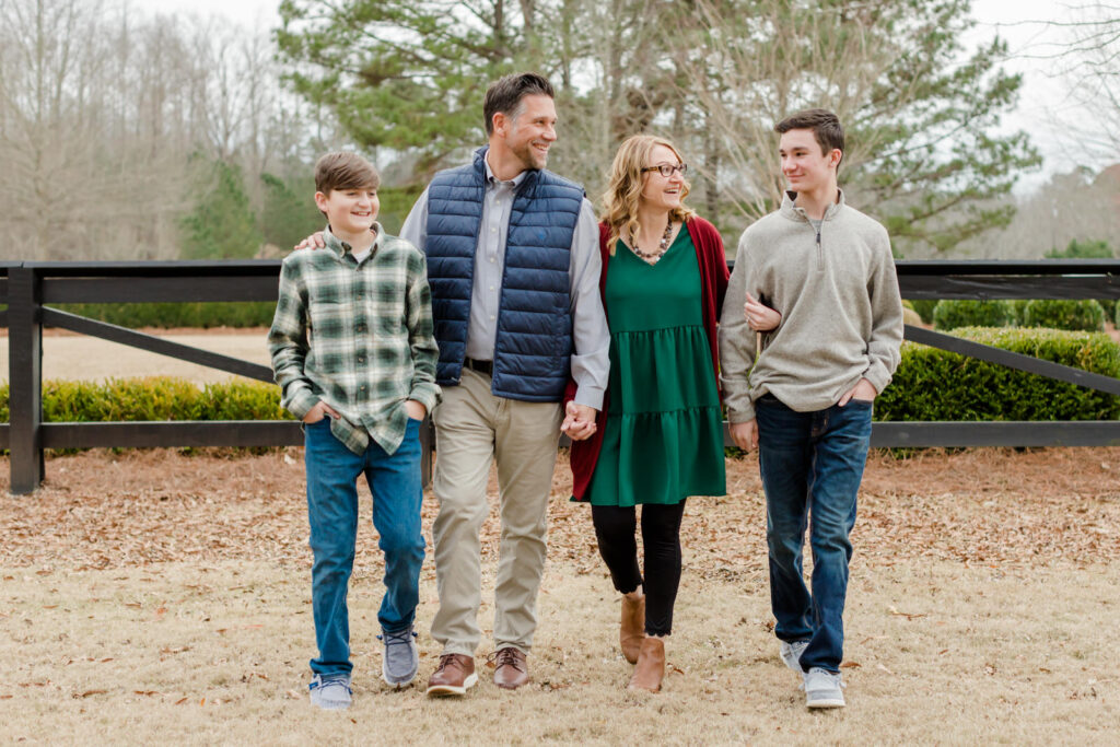 A family of 4 walk together smiling at each other with two teenage boys on the ends in Flowery Branch, Georgia at a mini-session.
