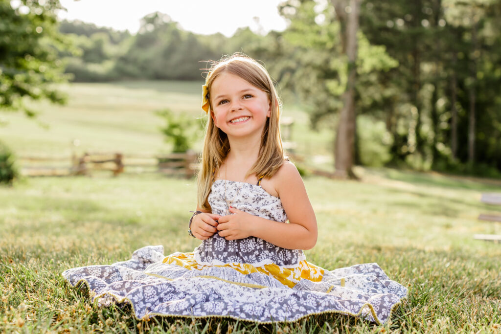 Little girl sits down and smiles at camera with her dress all around her in an open field. 