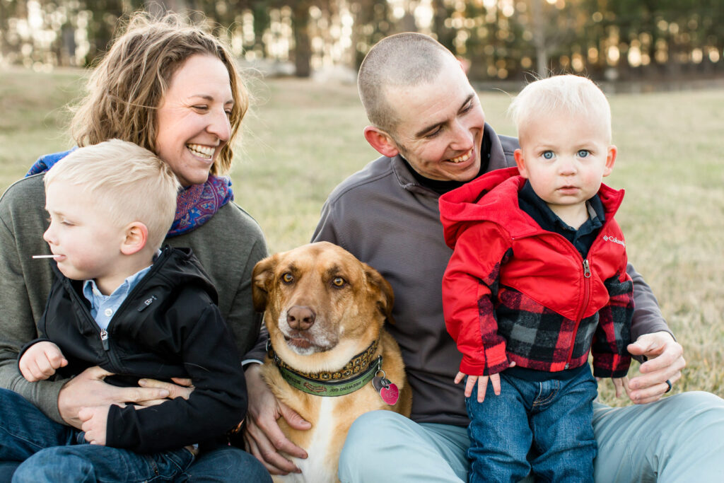 Parents hold their baby and toddler and smile at them while their dog cuddles between them during their family session with a dog in Flowery Branch
