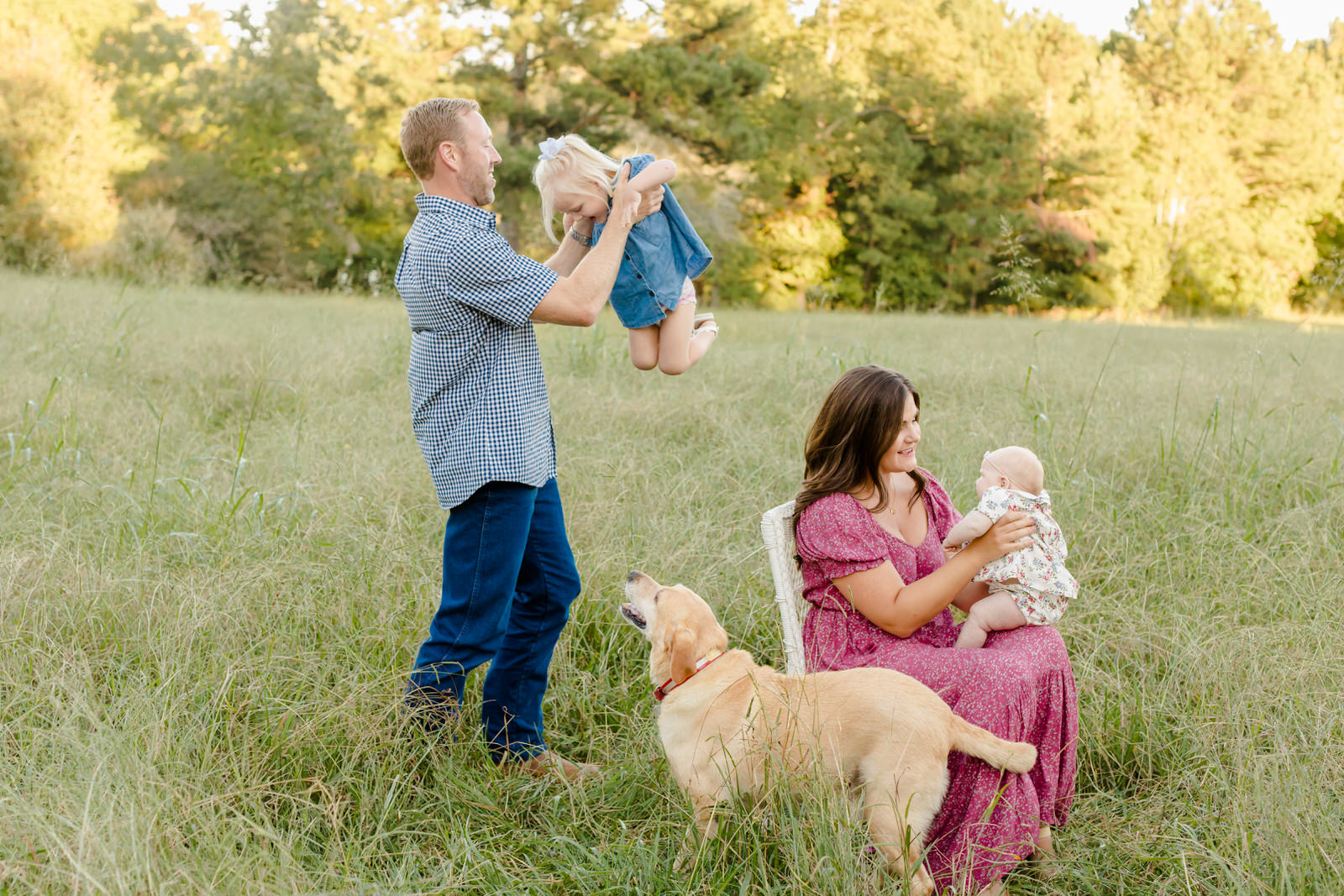 Mom sits in a field and smiles at her baby on her lap while Dad twirls his daughter around and dog looks on at Little Mulberry Park in Auburn, Georgia.