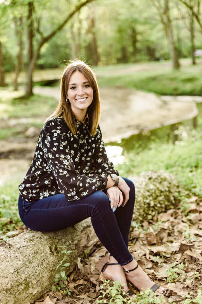 Senior girl sits on a log in front of a creek.