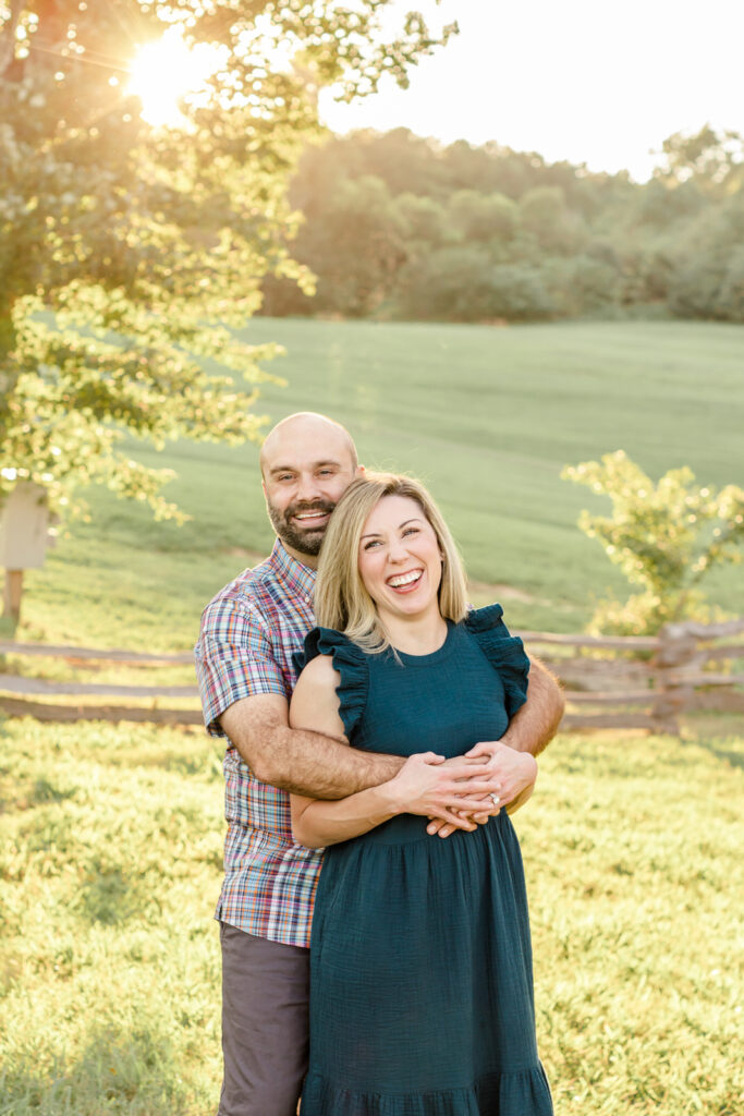 Husband wraps his arms around wife and they smile at camera in front of beautiful open field.