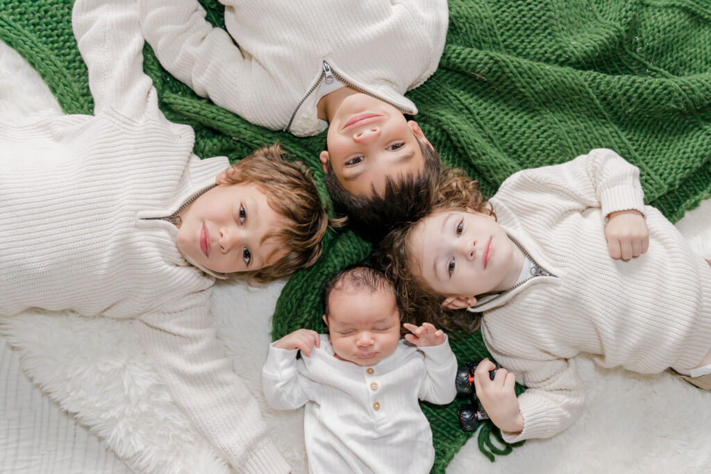 Four brothers lay on bed with heads touching and smile at camera during the newborn session for the youngest in Buford, GA