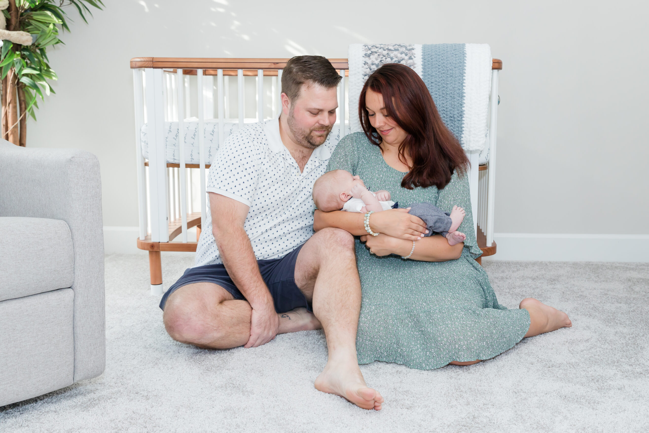Mom and Dad hold newborn baby in nursery and sit in front of crib. Mom wears a light green dress for the session.